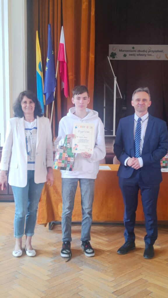 uczeń trzymający dyplom i nagrody stojący pomiędzy nauczycielami organizującymi konkurs
