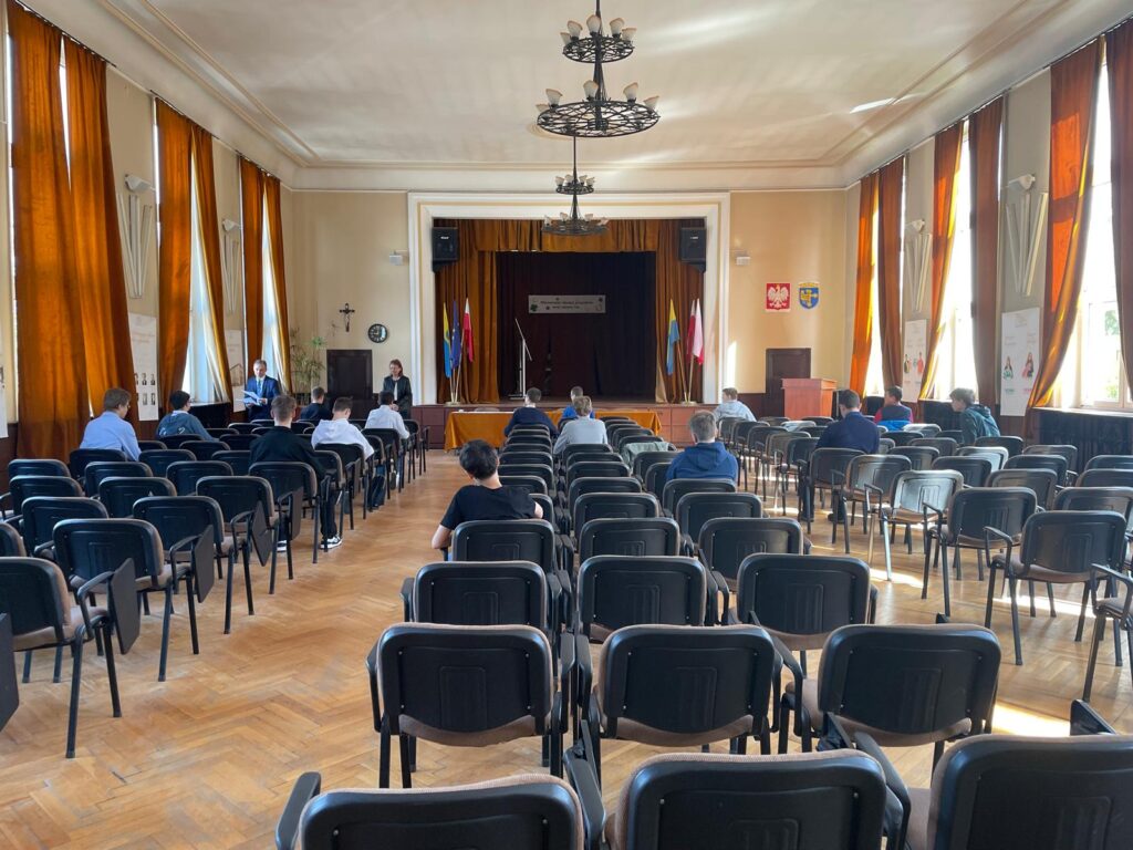 zdjęcie przedstawiające aule szkoły z widocznymi krzesłami, na krzesłach siedzący uczniowie w tle widać scenę auli szkoły