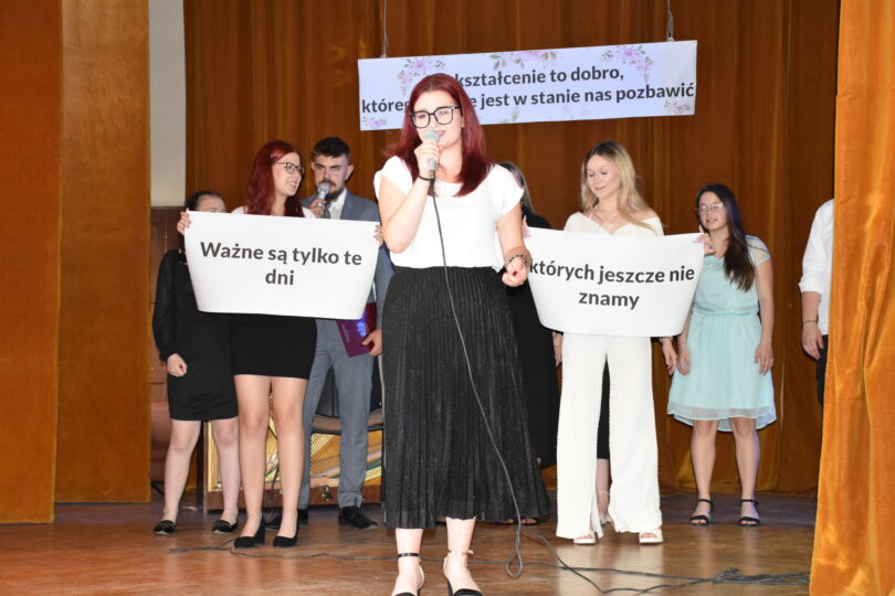 przedstawiona scena auli szkoły, na której uczennica śpiewa, w tle uczniowie trzymający napis ważne są te dni których nie znamy