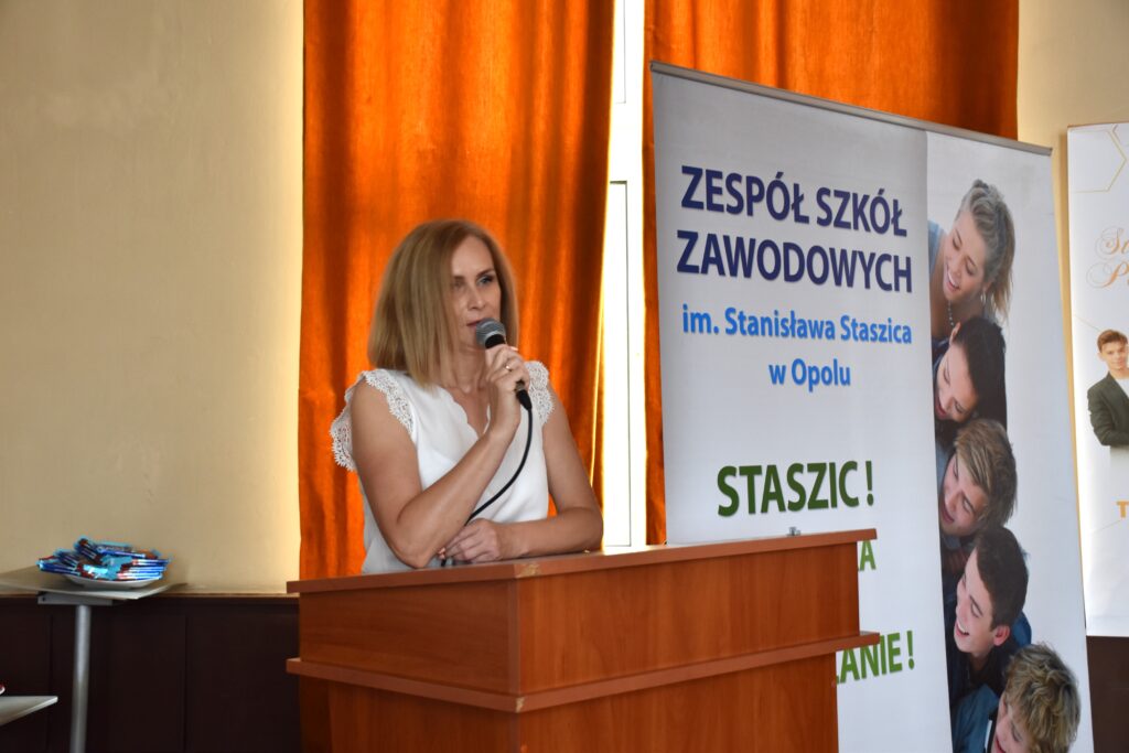 Pani dyrektor wygłasza przemówienie skierowane do uczniów, na tle plakatu Zespołu Szkół Zawodowych w Opolu