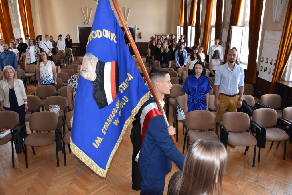 zdjęcie przedstawia poczet sztandarowy na tle społeczności uczniowskiej znajdującej się w auli